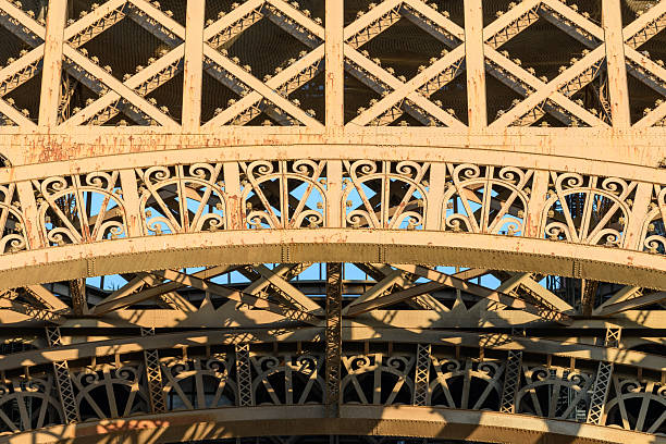 Detailed view of ornate iron arches and latticework on the Eiffel Tower, illuminated by warm sunset light and casting intricate shadows across the weathered metal surface.