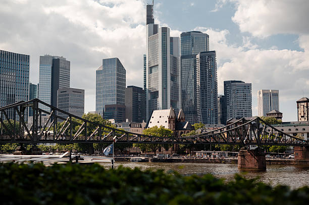 The Main River flows through Frankfurt, Germany, with the iconic skyline featuring modern high-rises and a historic iron bridge in the foreground