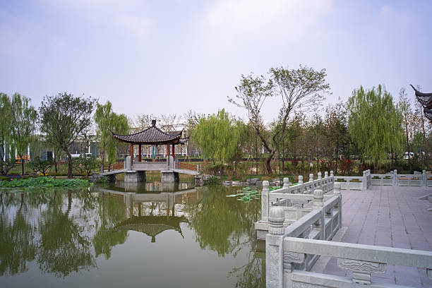 Wuhan, China - September 17, 2015: A peaceful scene featuring a traditional pavilion on a bridge reflecting in the water, framed by white stone railings at Wuhan Garden Expo Park.