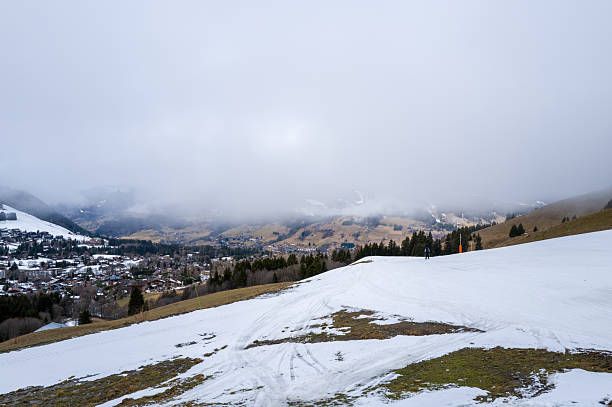 A ski slope with melting snow reveals grassy patches above the alpine village of Megeve, with low clouds and mist obscuring the distant mountains. The subdued winter light creates a tranquil, transitional atmosphere.