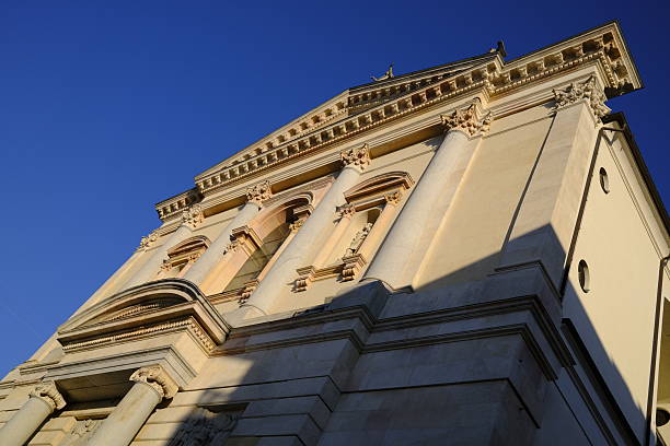 Low-angle view of a historic architectural facade with classical columns and decorative details, photographed in Gallarate, northern Italy. Warm sunlight highlights the stone surface against a deep blue sky, emphasizing symmetry, structure, and timeless design.