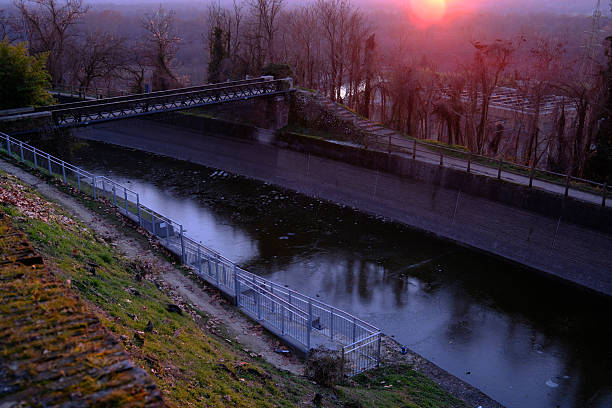 Sunset over a river landscape in Tornavento, northern Italy. The low sun casts warm light across the horizon, silhouetting trees and surrounding countryside, while reflections appear on the calm water below. The scene conveys tranquility, transition, and the peaceful atmosphere of the Italian landscape at dusk.