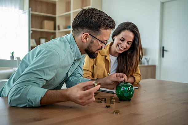 Happy couple saving coins into a green piggy bank on a wooden table, planning their financial future together in a cozy home setting with smiles and teamwork