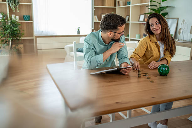 Young adult couple is making a budget and saving coins in a piggy bank for future financial planning, sitting at a wooden table in their bright living room