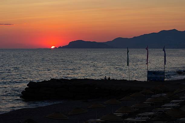 Peaceful sunset over the sea with mountain silhouettes, calm water and beach umbrellas in the foreground. Scenic coastal landscape at dusk, summer travel and relaxation mood