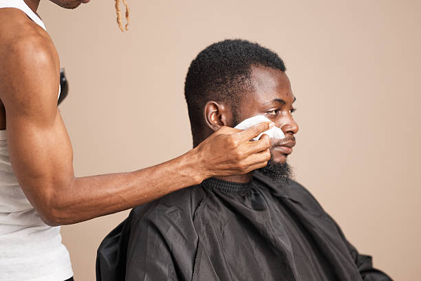 A young black man wearing a black barber cape sits calmly as a barber wipes his cheek with a cloth during a grooming session against a neutral studio background.
