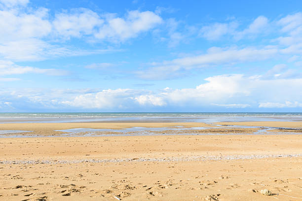 Wide open sandy beach at Sword Beach in Ouistreham stretches toward the calm sea under a bright blue sky with scattered clouds, creating a peaceful and inviting coastal scene.