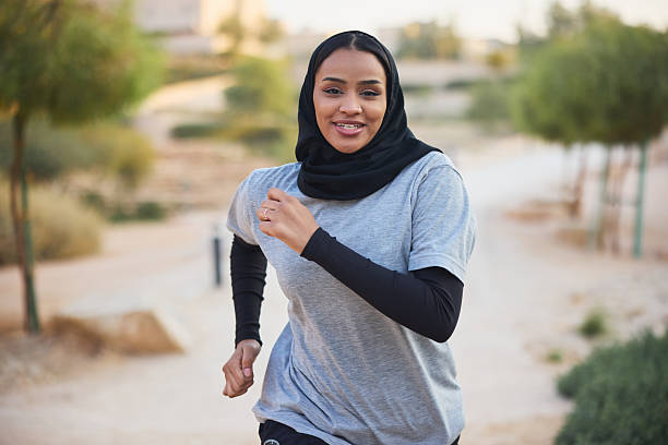 Determined young woman jogging in public park. Active female is wearing hijab. She is looking at camera.