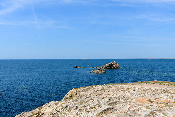 Jagged rocks extend into the deep blue ocean from a sunlit coastal outcrop at Pointe Beg-An-Aud in Saint Pierre Quiberon, with clear skies and tranquil water creating a peaceful seascape.