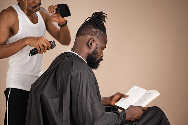 A barber trims the back of a bearded man’s head with clippers as the seated client with tied dreadlocks reads a book against a plain studio background.