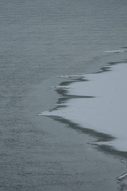 Frozen river or lake with visible ice layers and snowy banks. Dark water contrasts with white and pale blue ice, creating a clear boundary. Thin ice, snow patches, and some exposed sandy areas visible along the edge. The scene conveys a cold, winter atmosphere in a natural, isolated location.