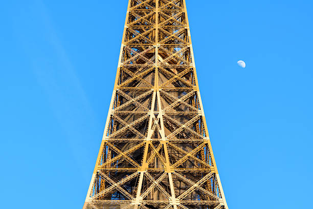 Upward view of the Eiffel Tower's golden iron lattice structure with a clear blue sky and the moon visible to the side, creating a striking geometric composition.