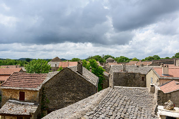 Panoramic view across weathered stone rooftops and old village houses in La Couvertoirade, with lush greenery and dramatic storm clouds overhead creating a moody, historic atmosphere.