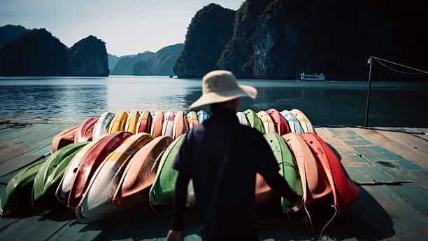 At the edge of Ha Long Bay, colorful kayaks rest beneath the wooden dock as a lone figure pauses before the journey. Beneath the beams, a large shadowy shape—often mistaken for a resting pet—adds a quiet sense of mystery, blending with the stillness of the bay and the calm rhythm of the morning.
