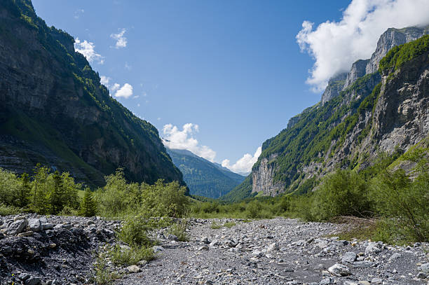 A broad rocky riverbed stretches between towering limestone cliffs and lush green slopes in Cirque du Fer-a-Cheval, under a bright blue sky with scattered clouds. Summer sunlight highlights the rugged textures and dramatic mountain scenery.