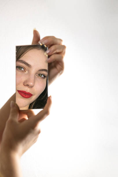 Conceptual portrait of a young woman with red lipstick looking into a mirror against a white background. The image symbolizes self-reflection, identity, inner beauty, emotional vulnerability, and modern minimalism