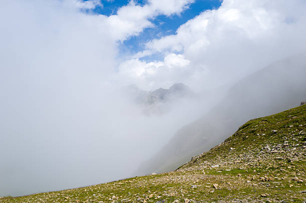Low clouds and mist drift across a stony green slope at Col du Bonhomme, partially obscuring distant mountain peaks under a bright patch of blue sky.