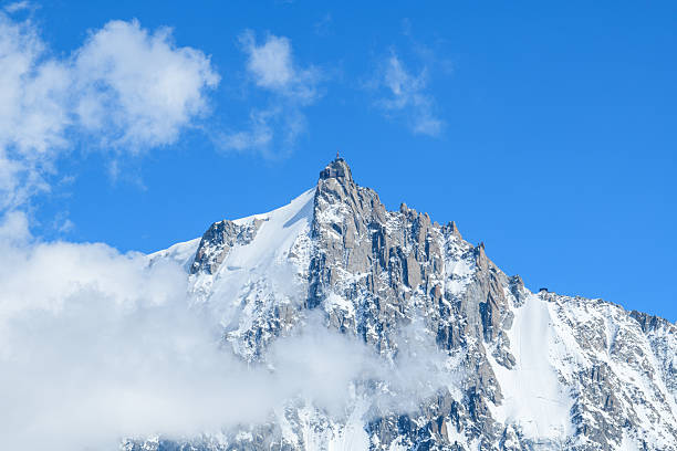 Aiguille du Midi stands tall with sharp, snow-covered ridges piercing through drifting clouds, set against a vivid blue sky in the French Alps.
