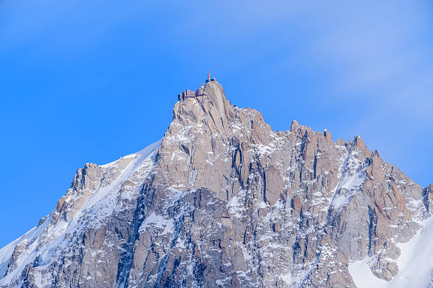 Sharp, rugged cliffs of Aiguille du Midi rise steeply, dusted with snow and illuminated by soft daylight beneath a vivid, cloud-streaked blue sky.