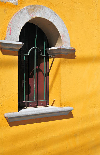 Quito, Pichincha, Ecuador: close-up architectural detail of a single ticket window at the Plaza de Toros. The window features a classic semi-circular arch with white stone trim and a gray stone sill set into a textured yellow stucco wall. Dark iron bars protect the dark wooden shutter behind. The high-contrast sunlight creates sharp shadows across the facade, emphasizing the depth and texture of the materials. This detail is representative of the traditional Spanish-influenced architecture used throughout the bullring's exterior. Bullfighting has been prohibited in the capital following a referendum, and the venue is now frequently used for 