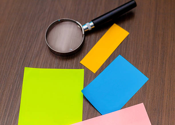 A magnifying glass with a black handle lies on a dark wood surface alongside scattered neon sticky notes in green, blue, yellow, and pink. The composition suggests research or an investigation.