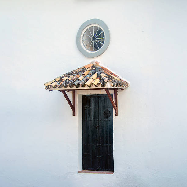 Minimalist architectural detail of a whitewashed wall featuring a small wooden door with a clay tile canopy and a circular window above, typical of an Andalusian white village in southern Spain.