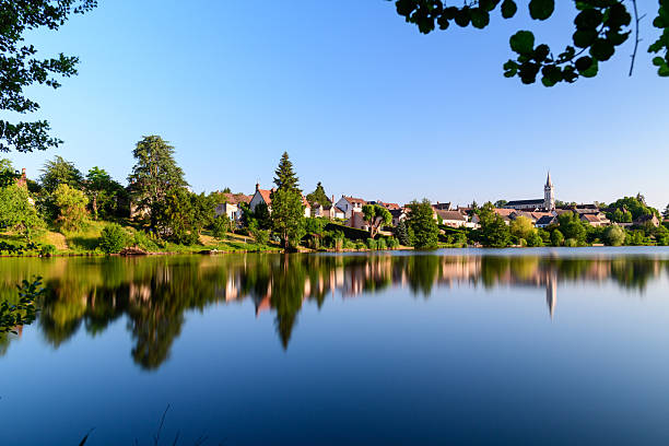 The calm waters of the pond reflect the rooftops, church spire, and vibrant greenery of Dampierre en Burly, framed by leafy branches under a cloudless blue sky. Crisp morning light and stillness create a harmonious rural scene.