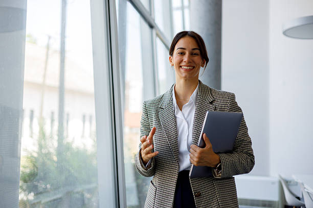 istock Confident businesswoman smiling, gesturing while holding a laptop, standing in a bright office with large windows 2255090974
