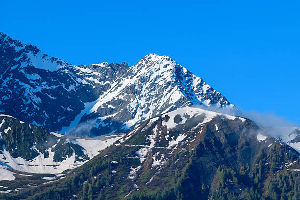 Dramatic view of Mont Lachat with its snow-dusted peak rising above steep, rocky slopes and scattered evergreen trees under a vivid blue sky. Crisp sunlight highlights the mountain's textures and late spring snow patches.