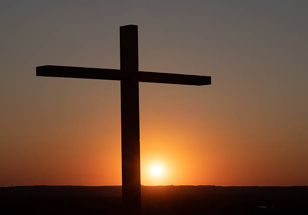 Silhouette of a tall black cross with the orange and yellow sun setting behind it is a symbol of Easter Resurrection