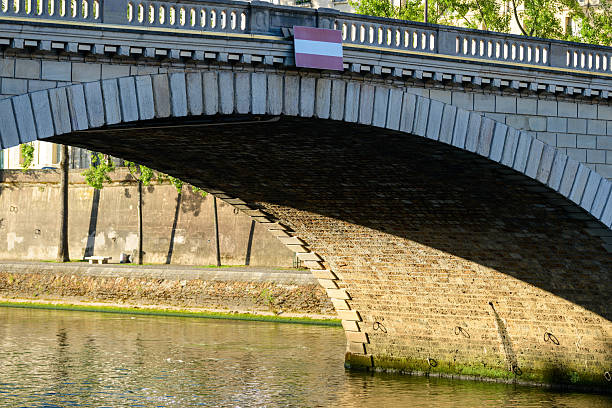 Detailed view of the underside of Le Pont Louis-Philippe in Paris, with sunlight and shadow highlighting the textured masonry above the calm Seine River.