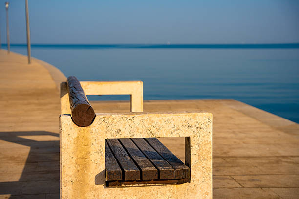 Porec, Croatia - August 13, 2025: Minimalist bench on a sunlit pier facing the calm sea, warm stone textures and soft morning light creating a serene coastal atmosphere.