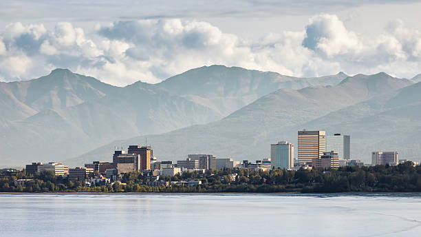 City skyline of Anchorage along the Cook Inlet with modern downtown buildings backed by the Chugach Mountains under soft daylight and scattered clouds. Iconic urban Alaska scene combining city life and dramatic mountain landscape, ideal for business, travel, and editorial use. Anchorage, Anchorage Municipality, Alaska, United States, North America