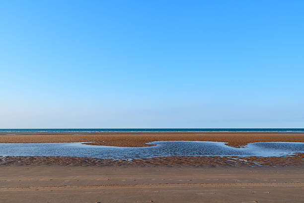 Expansive tidal flats with shallow water pools stretch across the sandy Omaha Beach under a vast, clear blue sky. The tranquil coastal scene is open and minimal, evoking calm and simplicity.