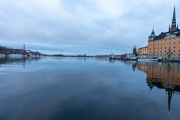 Calm winter reflections across the waterfront in Stockholm, Sweden, with the historic skyline of Gamla Stan and the soaring spire of Riddarholmen Church mirrored on the glassy harbor.