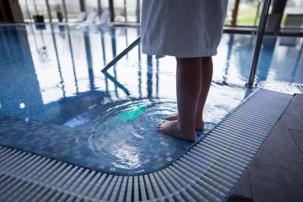 A young child stands at the edge of an indoor pool, toes in the water, wearing a light robe. Reflections shimmer across the tiled pool floor in a calm, family-friendly setting.