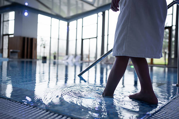 A person in a white robe walks barefoot through an indoor pool, creating soft reflections on the water. Captures calm wellness, leisure, and hotel spa vibes.