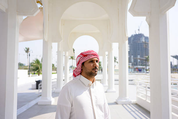 Saudi arabian man looking away while wearing traditional thobe and ghutra, standing outdoors under white archways representing Middle Eastern architecture and culture