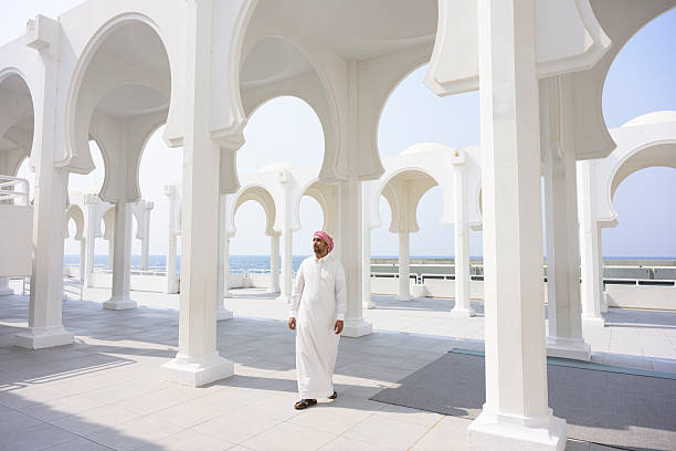 Middle Eastern man in white thobe and red ghutra walks beneath elegant mosque arches overlooking the clear blue sea, sunlight casting serene shadows on pillars and floor. Colonnade of arches adjacent to Al-Rahma  Mosque in Jeddah, Saudi Arabia.