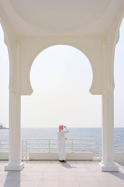 Mid adult Arab male in traditional white thobe and red ghutra, standing on a white tiled platform, looking out at the calm ocean through a modern archway