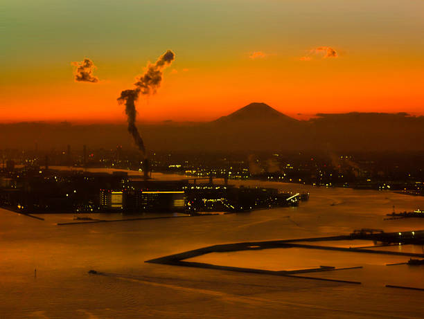 Tokyo Bay sunset aerial landscape with Fuji Mountain, industrial buildings, and ships in the harbor against a dramatic orangered sky over Haneda International Airport in Japan. A foggy soft focus image.