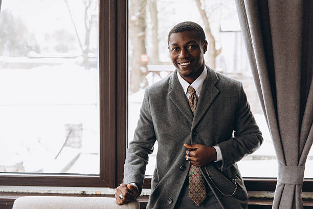 Smiling African American businessman looking at camera, standing by window in a suit and overcoat. Professionals and success concept