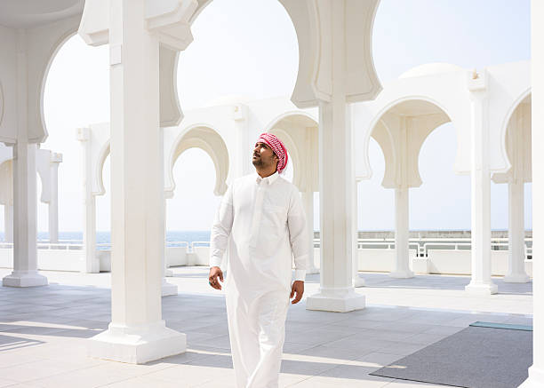 Arab man wearing traditional thobe and ghutra walking through modern white Islamic architecture with arches, observing the clear sky above and ocean in the background