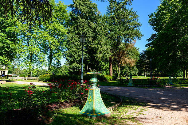 A lush landscaped park in Saint-Honore-les-Bains features a decorative green lamp post, blooming red rose bushes, and tall leafy trees under bright summer sunlight.
