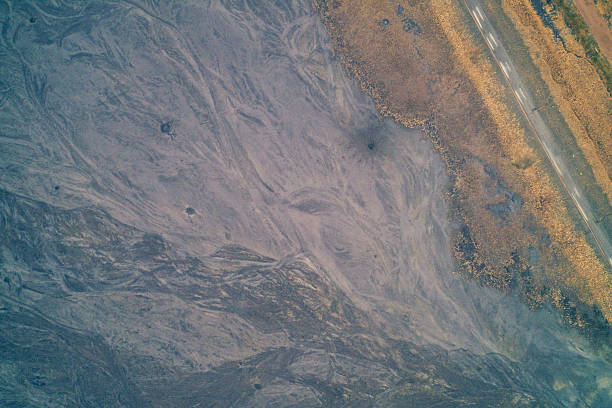 Aerial top view of a dried industrial pond bottom revealing grey toxic waste patterns next to a road and withered grass. A dystopian landscape of environmental damage, ash dump residue, and water scarcity consequences.