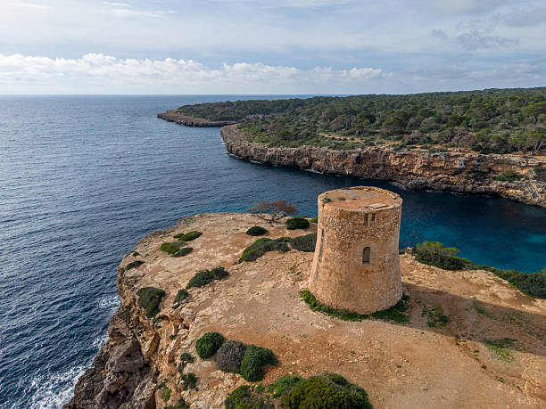 Aerial view of the historic stone watchtower at Cala Pi on the island of Mallorca, Spain. The old coastal tower stands on rugged cliffs above the turquoise Mediterranean Sea, surrounded by wild coastal vegetation and dramatic rocky coastline under a partly cloudy sky. A scenic and iconic Balearic landscape.