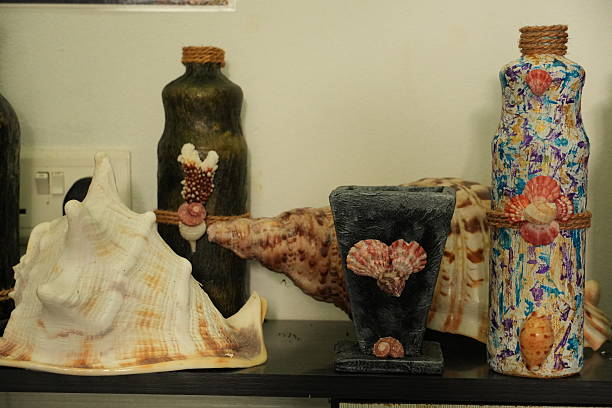 A collection of handmade sea-themed decorative items, including shell-decorated bottles and a conch shell, arranged on a shelf.