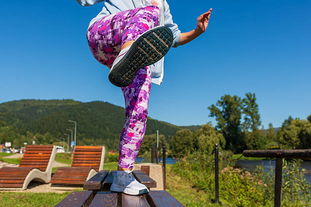 Low angle view of an active woman doing a high kick balance exercise on a wooden picnic table near a river, with green hills and a clear blue sky behind. Dynamic motion, strength, stretching, and outdoor training concept