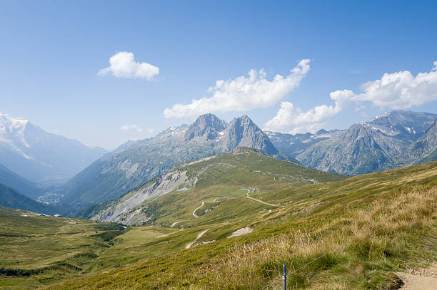 Jagged mountain summits rise above rolling green meadows and winding trails in a wide alpine valley under a bright blue sky with scattered clouds.