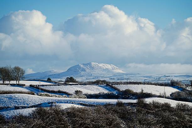Winter snow scene near Ballymena with Slemish mountain in the distance, and the rolling hills, grass fields and hedges of the County Antrim countryside, Northern Ireland. Shot on a sunny day with blue sky and snow clouds. It is a famous place because St Patrick herded sheep on Slemish mountain in his early life. The mountain is the remains of an extinct volcano and attracts many tourists.
